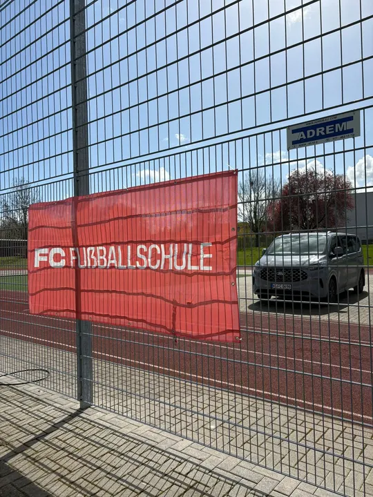 Banner der FC Fußballschule am Zaun zwischen Laufbahn und Parkplatz