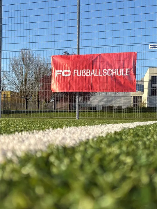 Banner der FC Fußballschule am Zaun des Sportplatzes vor blauem Himmel