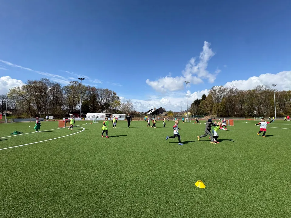 Weite Aufnahme des Kunstrasenplatzes mit vielen Kindern in Spielformen bei Sonnenschein