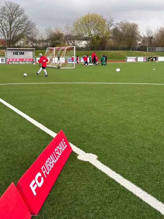 Rote Bande der FC Fußballschule am Spielfeldrand, dahinter Kinder beim Training am Tor