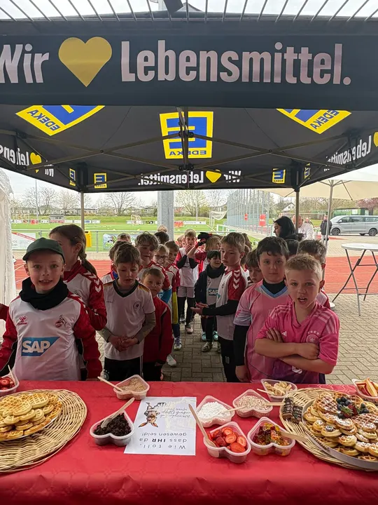 Kinder stehen unter dem EDEKA-Pavillon vor dem Tisch mit Waffeln und frischem Obst an