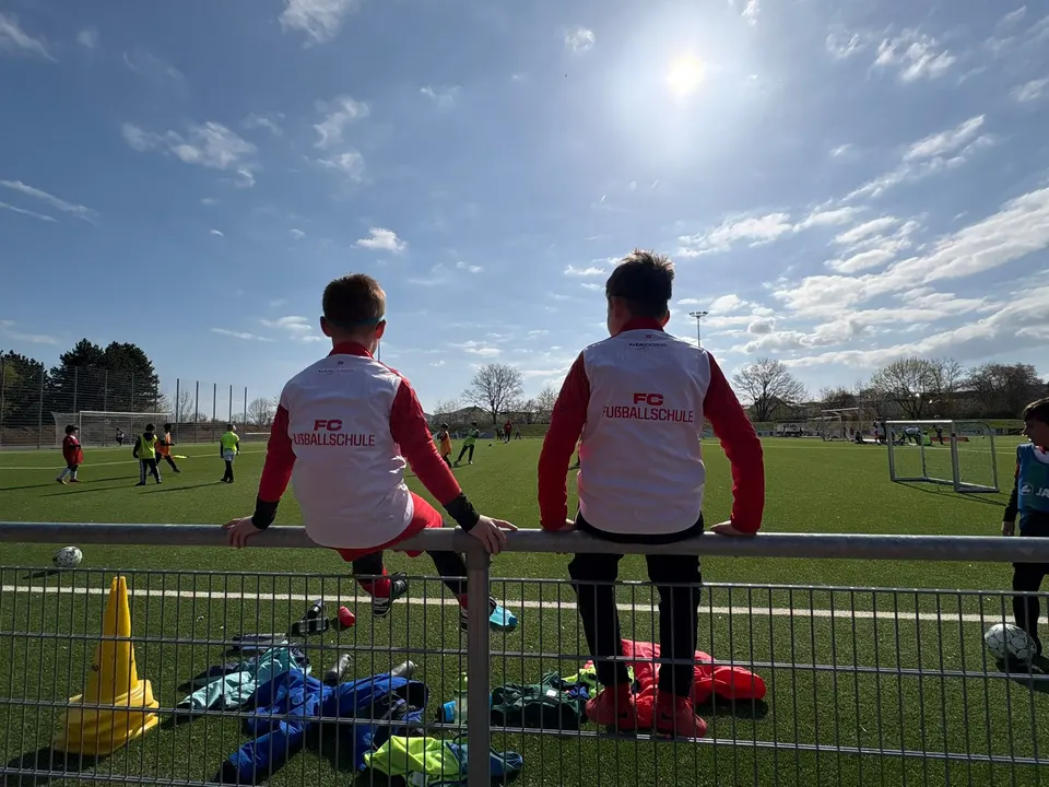 Zwei Jungen in Leibchen der FC Fußballschule sitzen auf dem Geländer und sehen dem Training zu